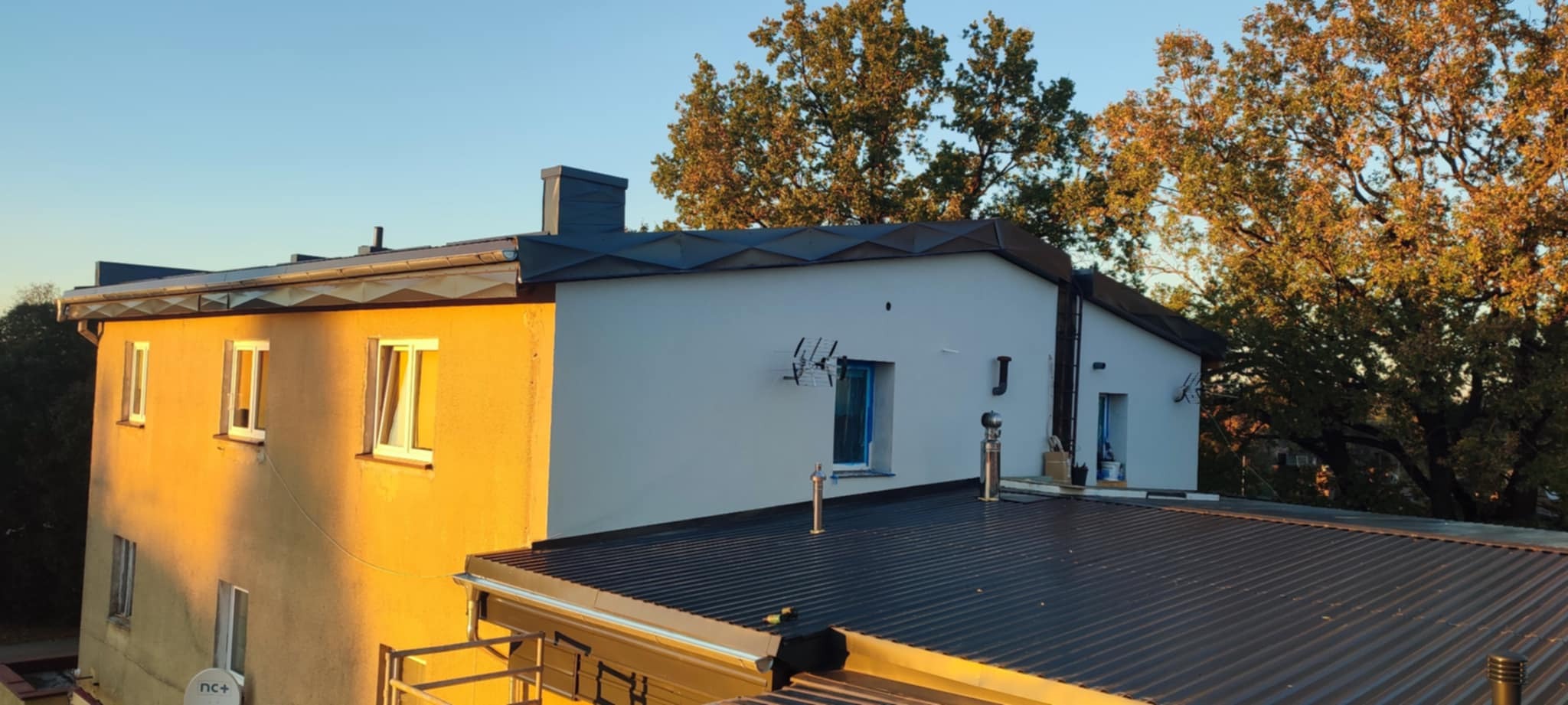 Nowy, ciemny dach z blachy trapezowej na dwukondygnacyjnym budynku z żółtą i białą elewacją, widoczne kominy i anteny. W tle drzewa i błękitne niebo.