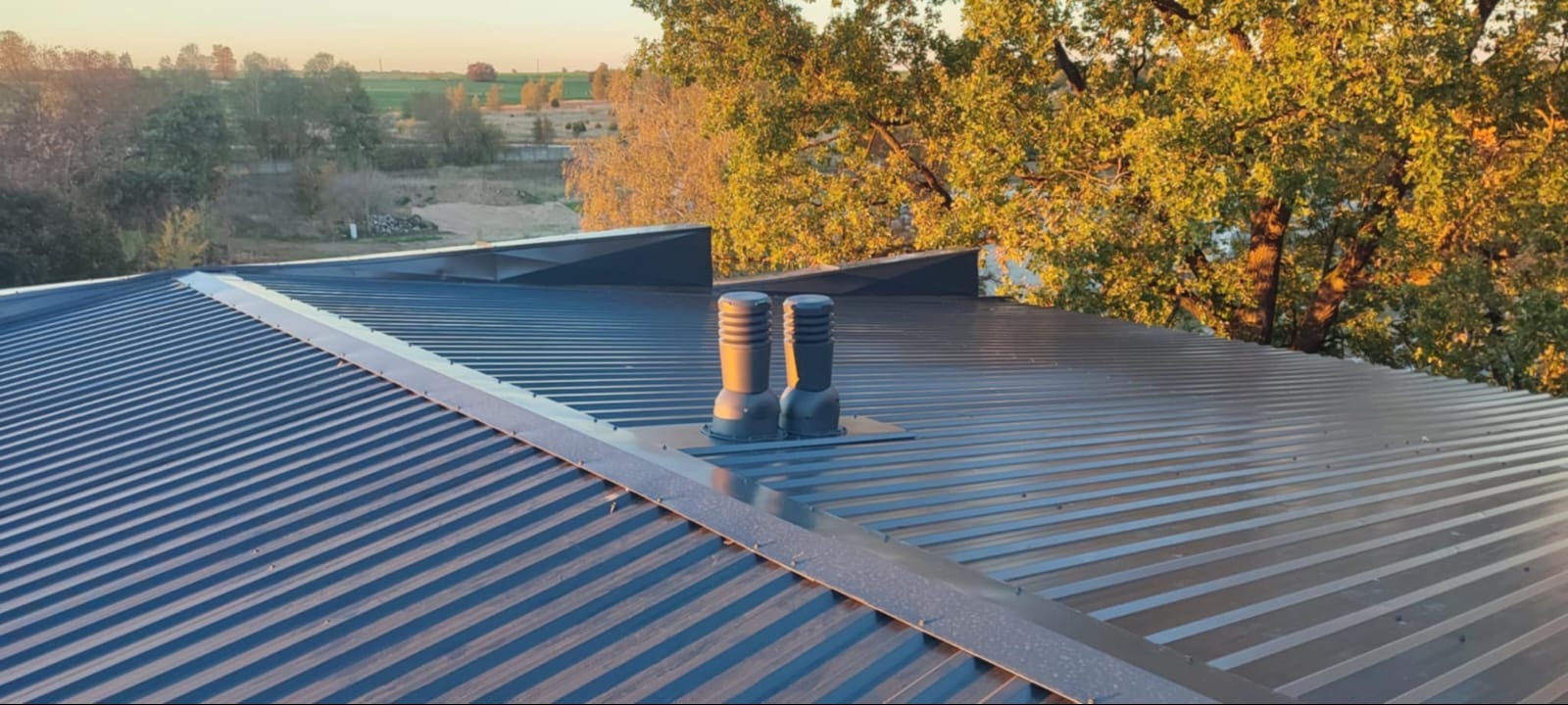 Nowy, blaszany dach w kolorze grafitowym z dwoma kominkami wentylacyjnymi na tle drzew w jesiennej scenerii, widziany z góry.