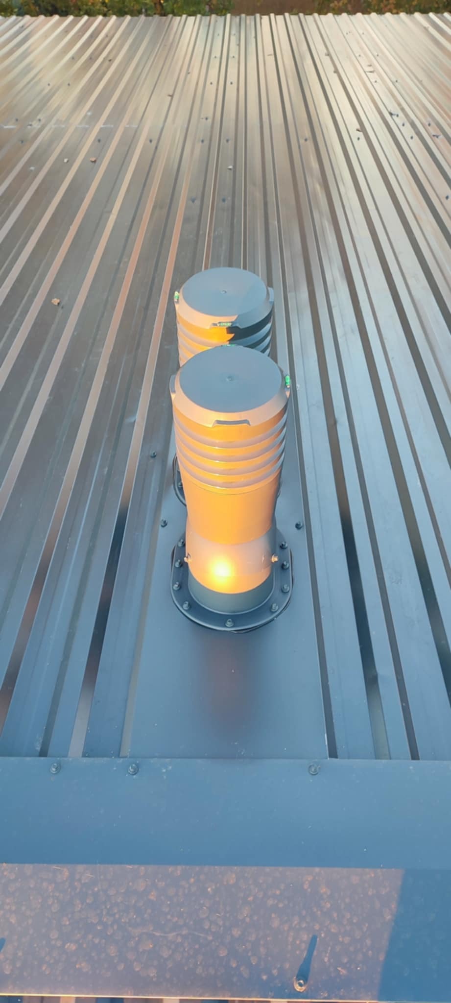 Metalowy dach z dwoma kominami wentylacyjnymi, widok z góry. Komin z pomarańczowym światłem wewnątrz. Dach pokryty blachą trapezową w odcieniach szarości.