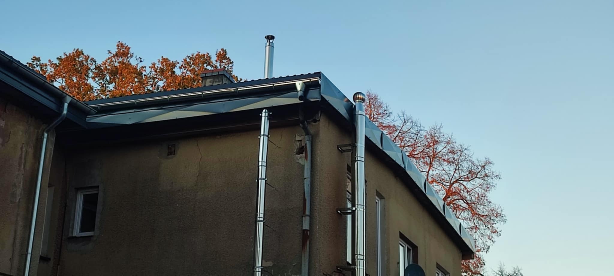 Szary budynek z nowym, ciemnym dachem i błyszczącymi, metalowymi rynnami oraz kominami na tle błękitnego nieba i drzew z pomarańczowymi liśćmi.