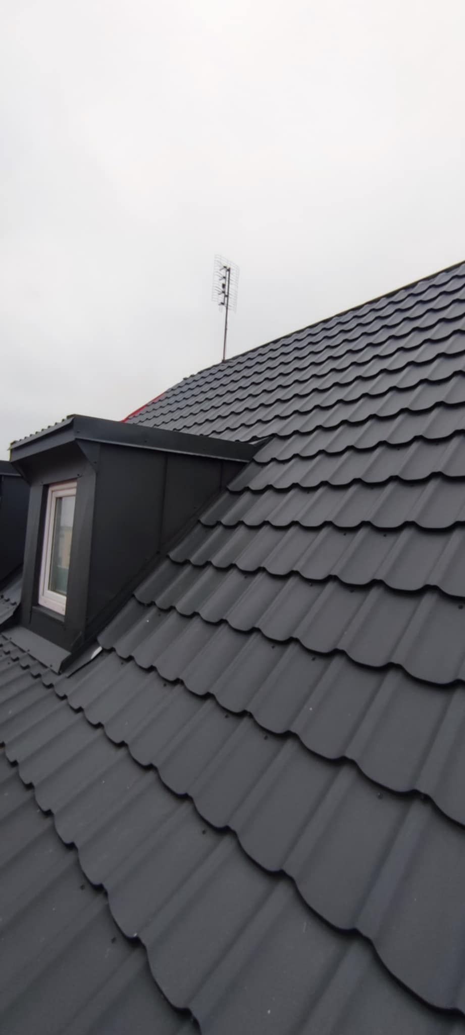 Grafitowy dach z blachy trapezowej z oknem dachowym i anteną telewizyjną na tle pochmurnego nieba. Widok z góry.