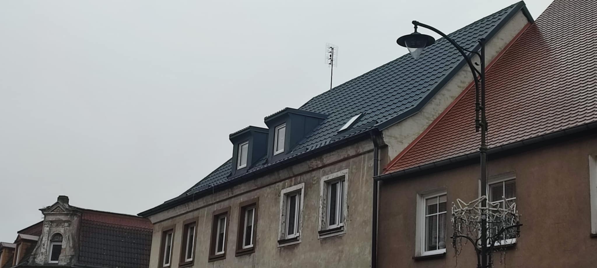 Szary dach z lukarnami na budynku z jasną elewacją, widoczna antena i latarnia uliczna. Dachówka w odcieniach szarości, budynek sąsiaduje z innym o czerwonym dachu.