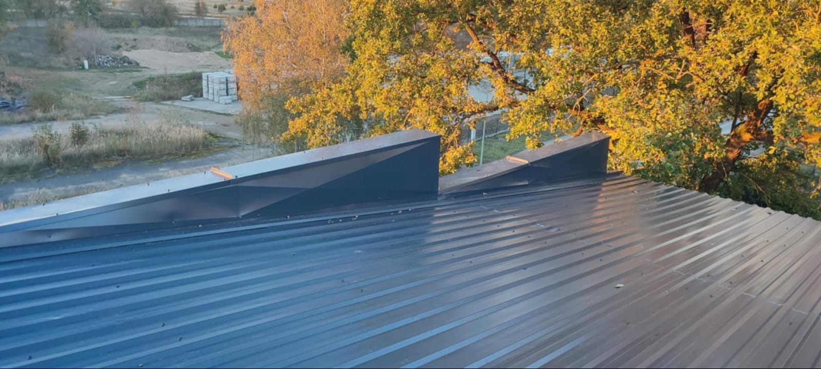 Szary dach z blachy trapezowej z obróbkami blacharskimi w kształcie kominów wentylacyjnych, na tle drzew i budynków w oddali, w słoneczny dzień.