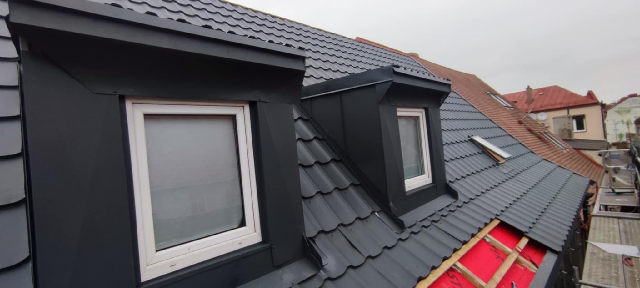 Czarny dach z oknami dachowymi i obróbkami blacharskimi, widok z góry, fragment czerwonej membrany dachowej, sąsiedni dach z czerwoną dachówką w tle.