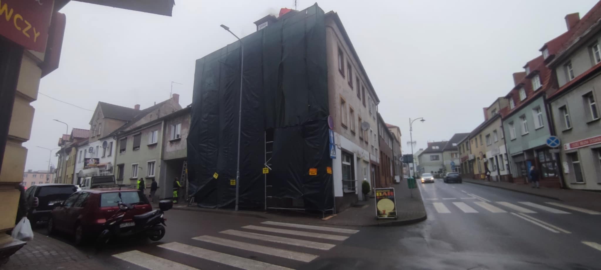 Budynek w rusztowaniach, osłonięty czarną folią, prawdopodobnie w trakcie prac remontowych. Widoczne fragmenty dachu i elewacji. Ulica miejska, pochmurny dzień.