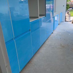 Ariel Borzymowski - Błękitne, lakierowane fronty szafek kuchennych na wymiar w trakcie montażu, widoczne puste przestrzenie na półki i szuflady, surowy beton na podłodze, okno w tle.