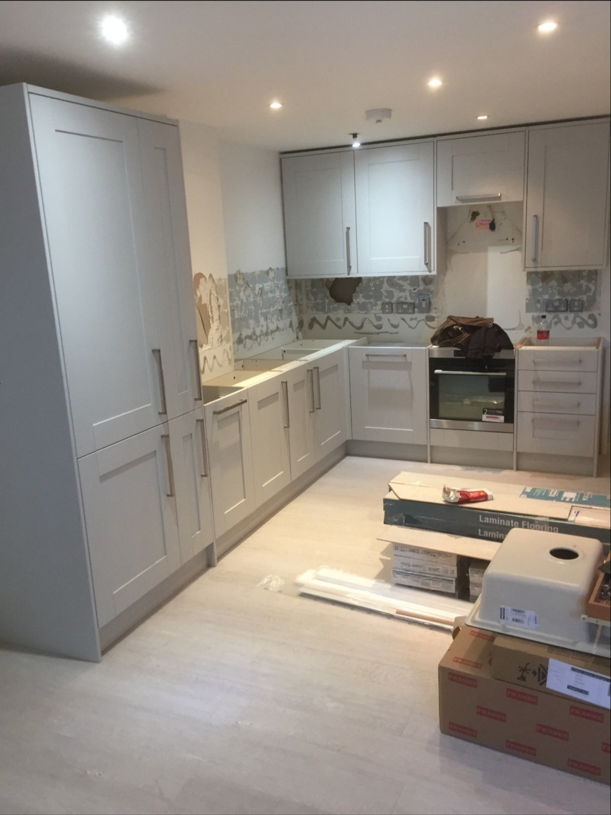 Montaż jasnoszarej kuchni na wymiar w trakcie: widoczne szafki, płyta grzewcza, kartony z panelami podłogowymi i zlewem. Ściany częściowo przygotowane pod płytki.