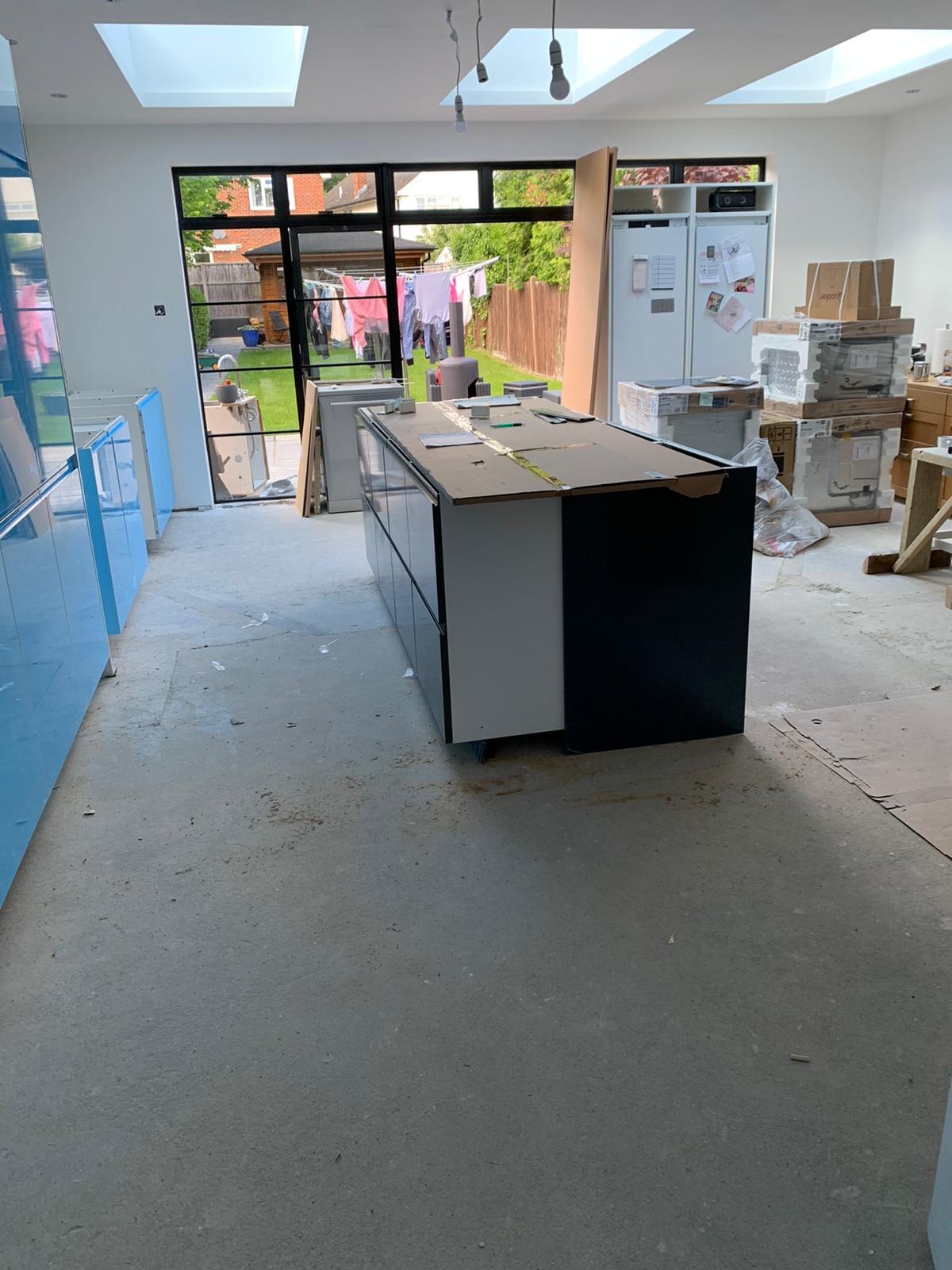 Montaż wyspy kuchennej z czarnymi i białymi frontami w surowym wnętrzu. Widoczne niebieskie szafki, okno z widokiem na ogród i kartony z wyposażeniem.