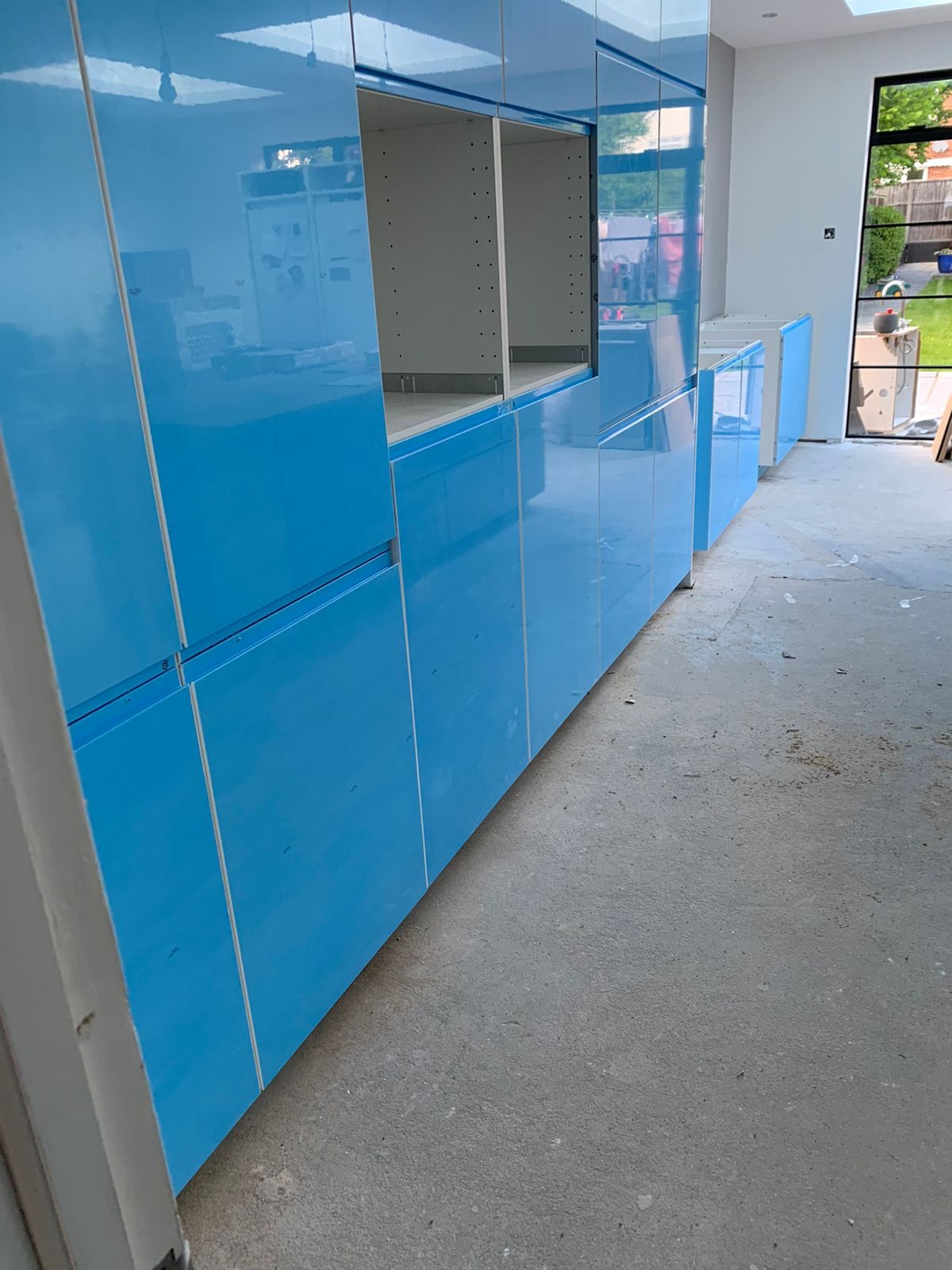 Błękitne, lakierowane fronty szafek kuchennych na wymiar w trakcie montażu, widoczne puste przestrzenie na półki i szuflady, surowy beton na podłodze, okno w tle.
