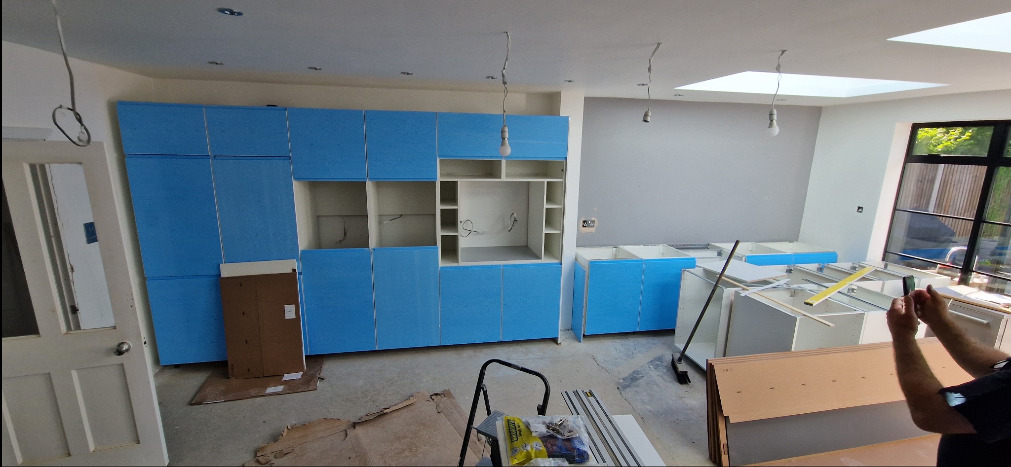 Niebieskie szafki kuchenne w trakcie montażu, widoczne elementy konstrukcyjne i narzędzia. Ujęcie z perspektywy osoby robiącej zdjęcie telefonem.