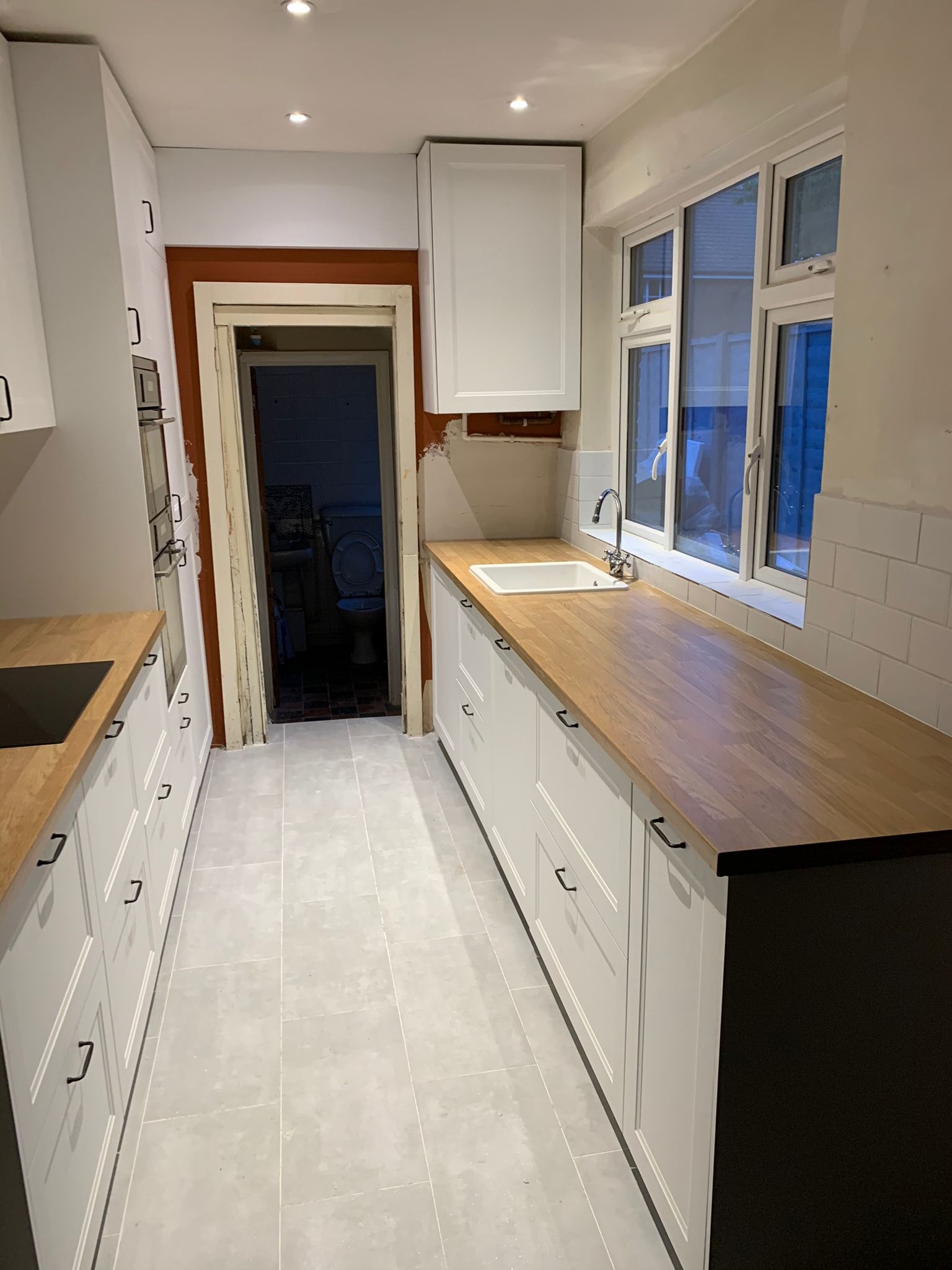Wąska kuchnia z białymi szafkami, drewnianym blatem i szarą podłogą. Widok na otwarte drzwi do łazienki. Umywalka przy oknie.