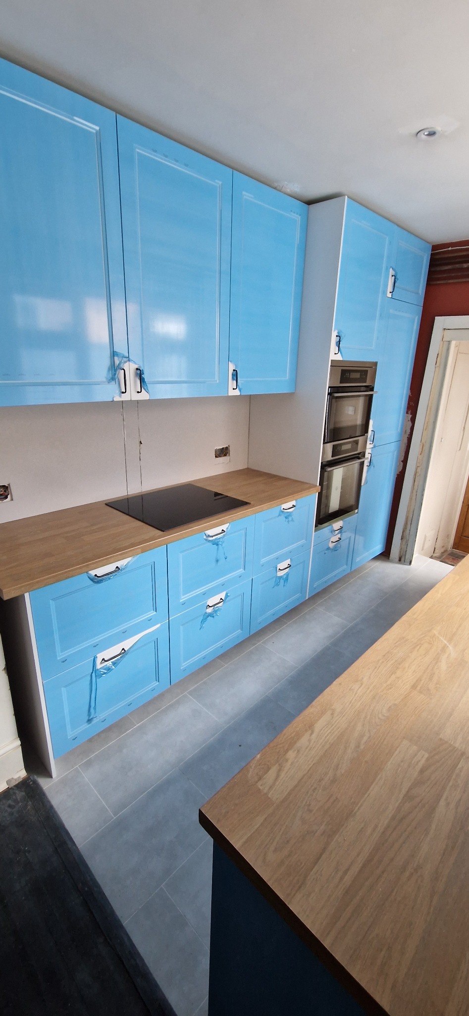 Niebieska zabudowa kuchenna z drewnianym blatem i płytą indukcyjną. Fronty szafek zabezpieczone folią ochronną. Szare płytki podłogowe. Widoczny fragment wyspy kuchennej.