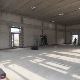 ROMKI Norbert Romek - Surowy stan wnętrza budynku z murem z betonu komórkowego, w tle widoczna osoba przy oknie, na podłodze narzędzia i skrzynki. Wnętrze w trakcie budowy.