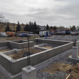 ROMKI Norbert Romek - Zalane betonem fundamenty domu jednorodzinnego z wystającymi prętami zbrojeniowymi, na tle plac budowy z maszynami i budynkami mieszkalnymi.