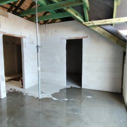ROMKI Norbert Romek - Wnętrze w trakcie murowania ścian z betonu komórkowego, widoczne drewniane elementy konstrukcji dachu i mokra posadzka. Podpora stropowa w centrum kadru.