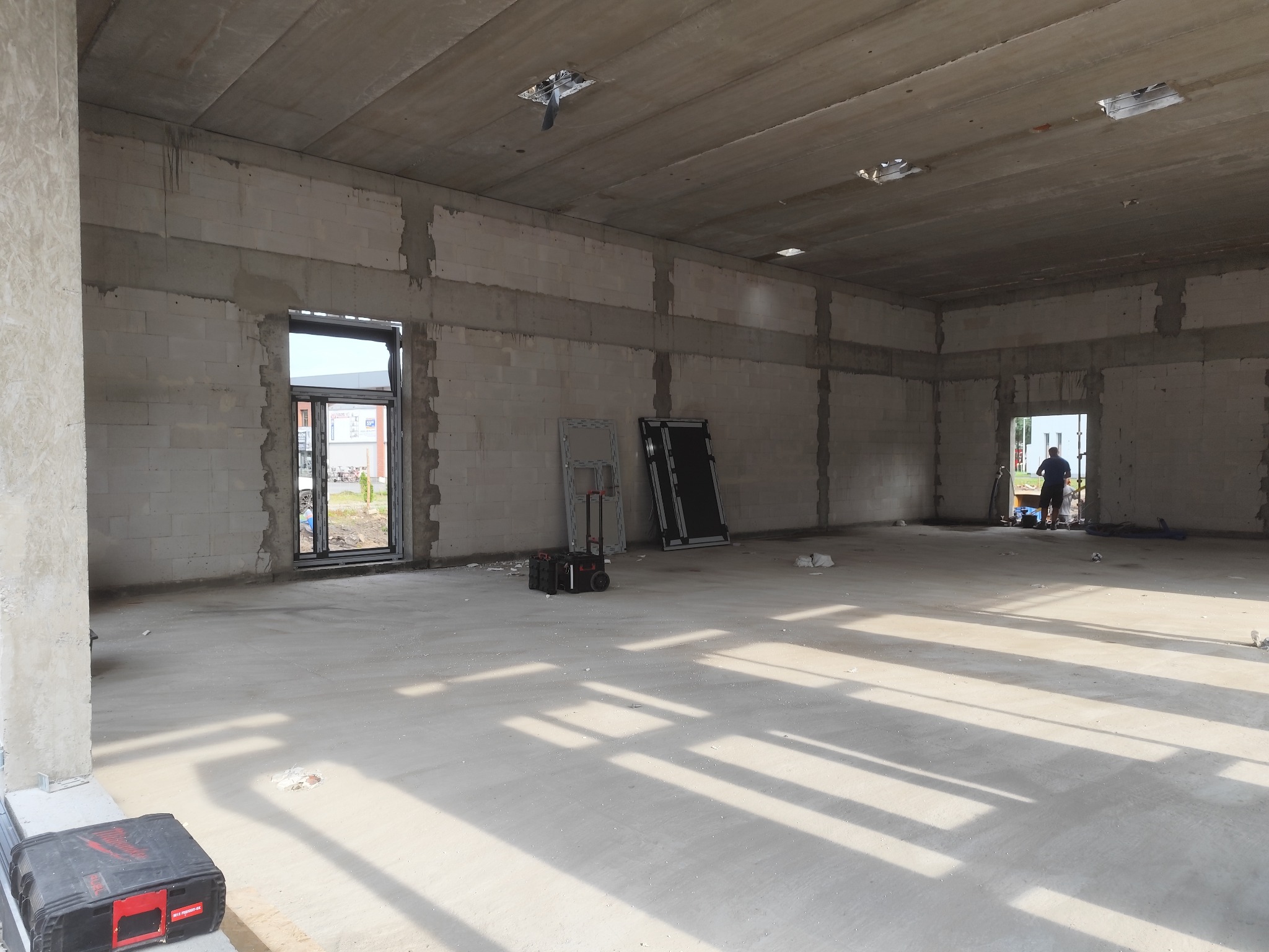 Surowy stan wnętrza budynku z murem z betonu komórkowego, w tle widoczna osoba przy oknie, na podłodze narzędzia i skrzynki. Wnętrze w trakcie budowy.