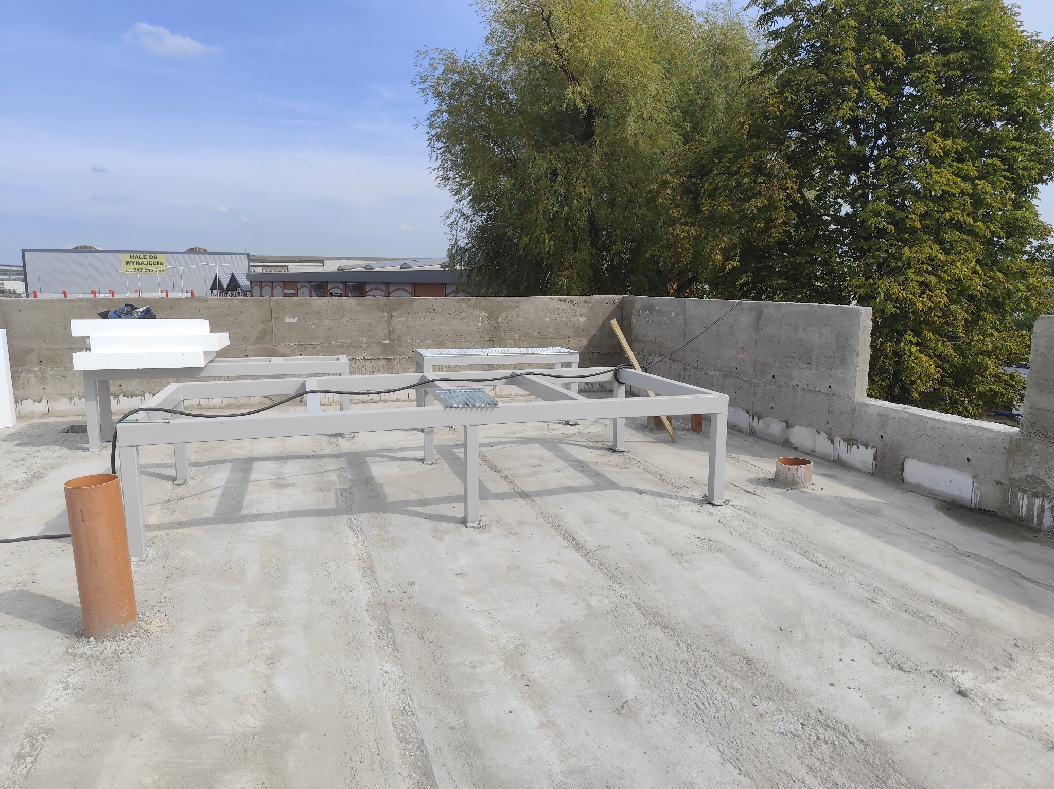 Betonowy dach z metalową konstrukcją pod panele słoneczne, widoczne elementy izolacji i fragment betonowej ściany. W tle drzewa i budynki.