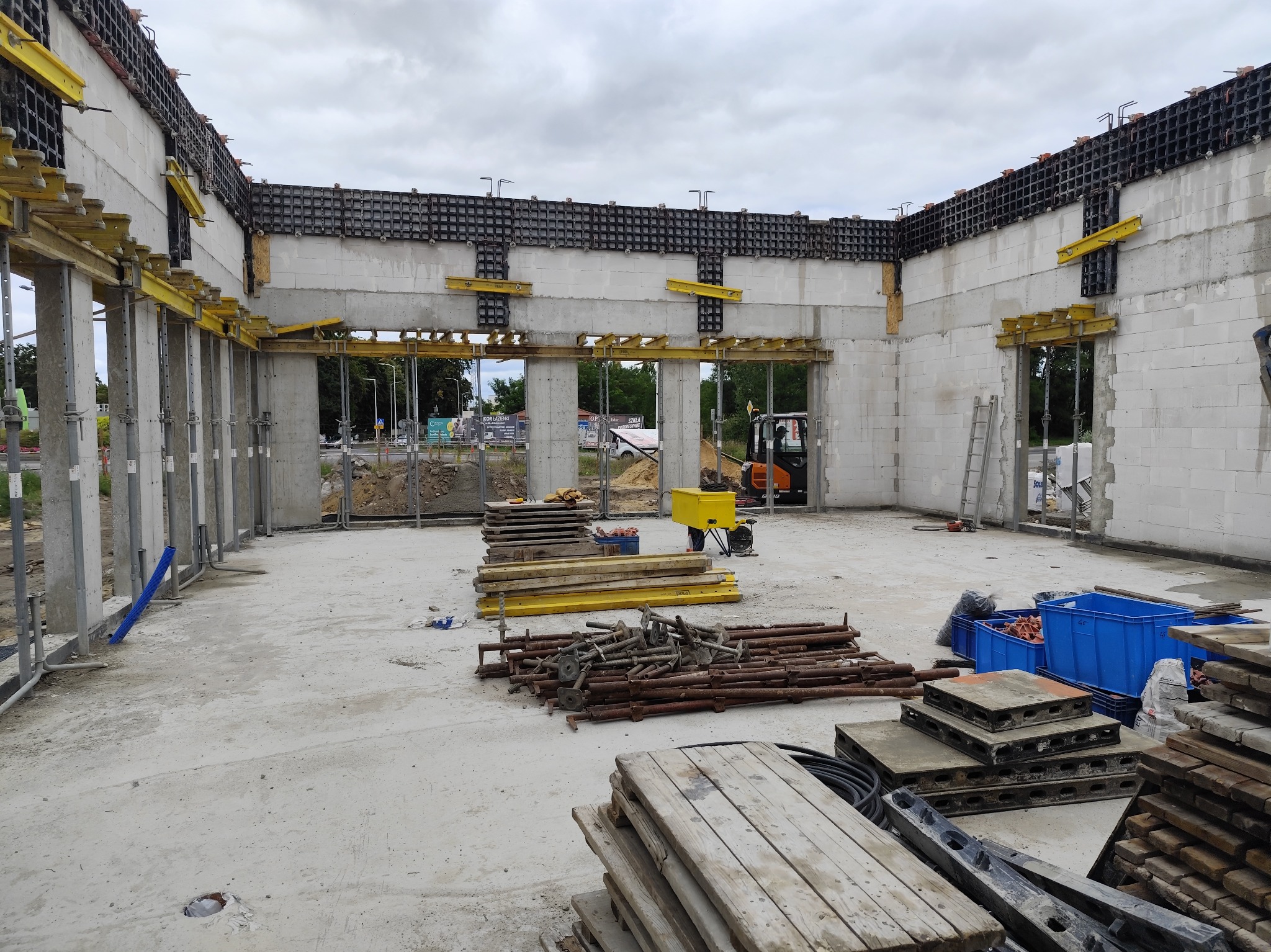 Wnętrze budynku w trakcie budowy z widocznymi ścianami z betonu komórkowego, szalunkami, podporami i sprzętem budowlanym, w tym małą koparką w tle.