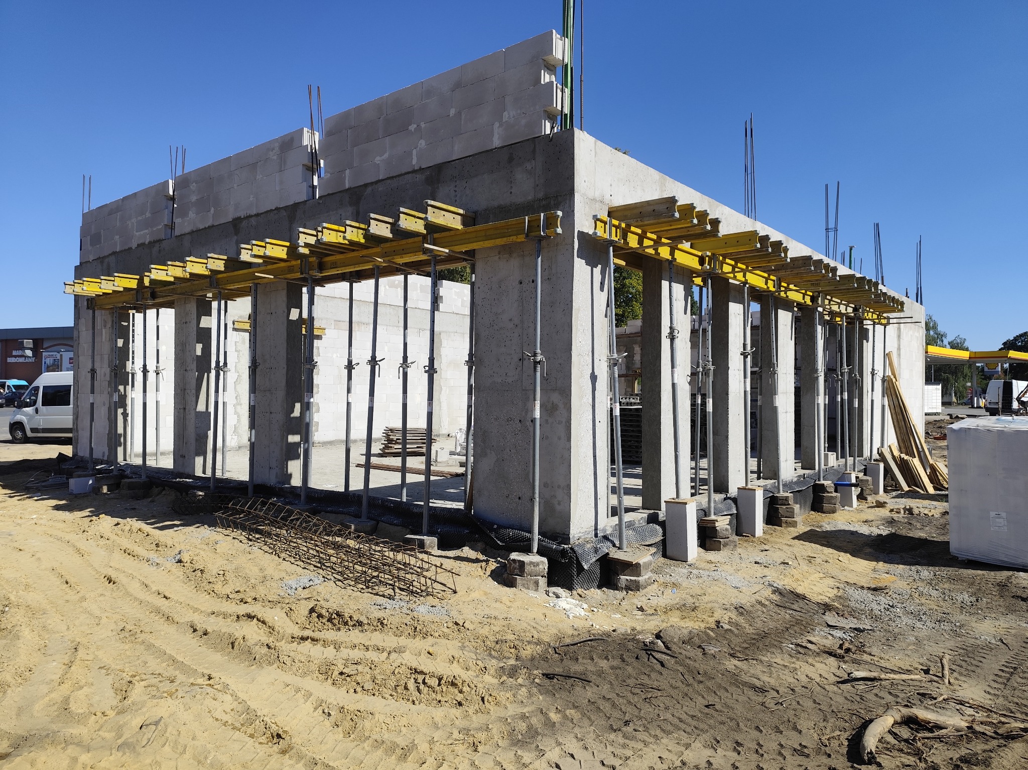 Budowa domu w stanie surowym otwartym: betonowe ściany, żółte podpory, widoczny teren budowy z piaskiem i elementami zbrojenia. Słoneczny dzień.