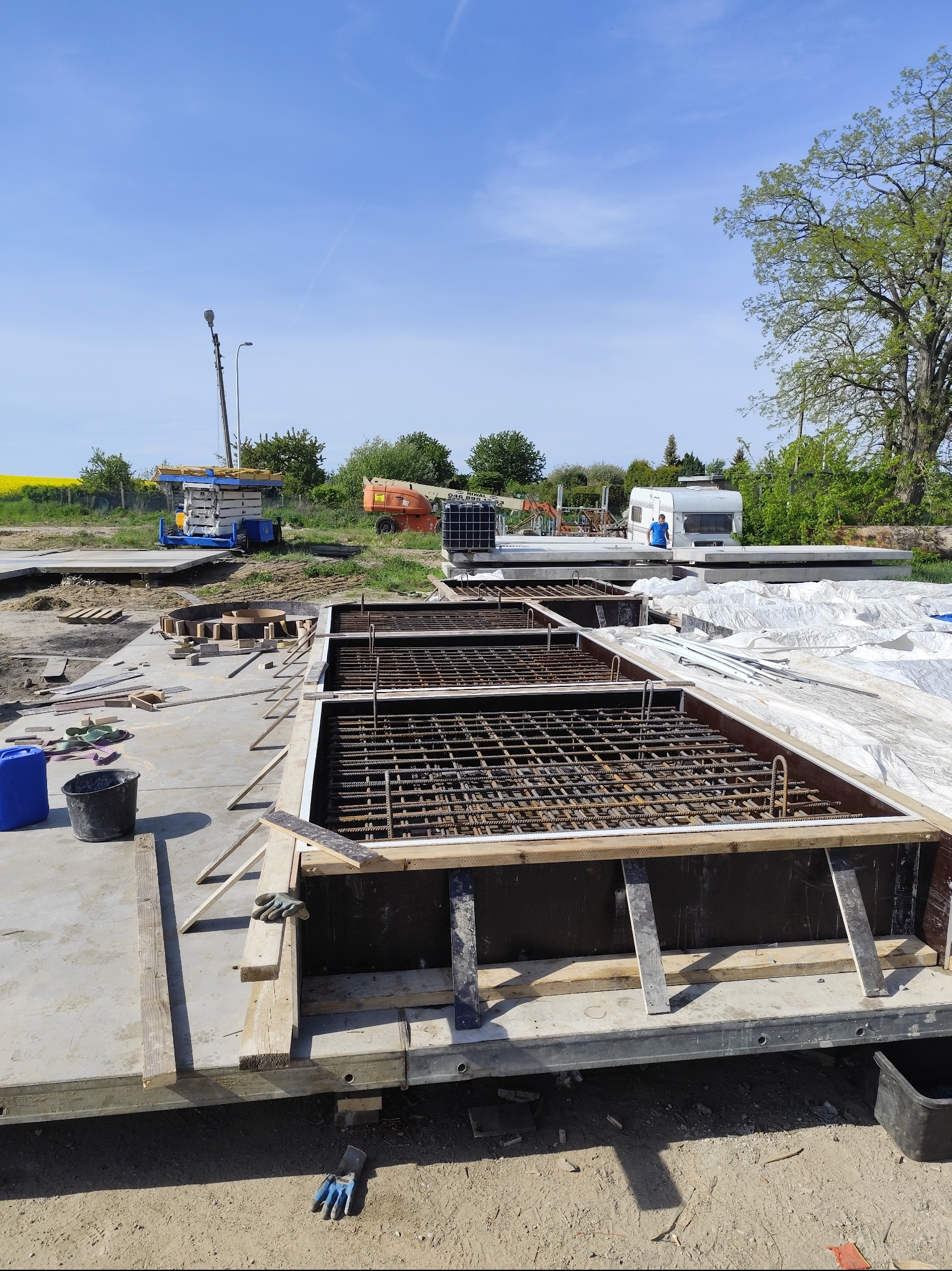 Przygotowanie do wylewania fundamentów: szalunek zbrojony stalą, widoczne maszyny budowlane w tle, błękitne niebo i zieleń drzew. Prace ziemne w toku.