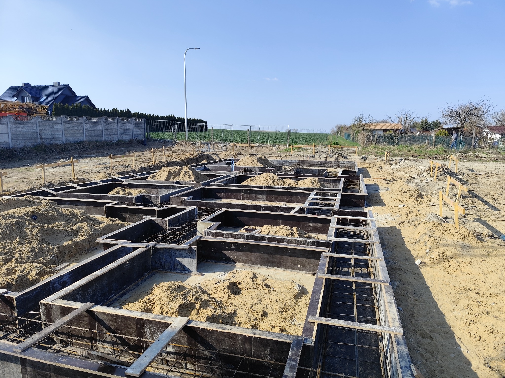 Szeroki kadr na przygotowane szalunki fundamentowe z widocznym zbrojeniem i częściowo zasypanym piaskiem, na tle krajobrazu z domem i polem w słoneczny dzień.