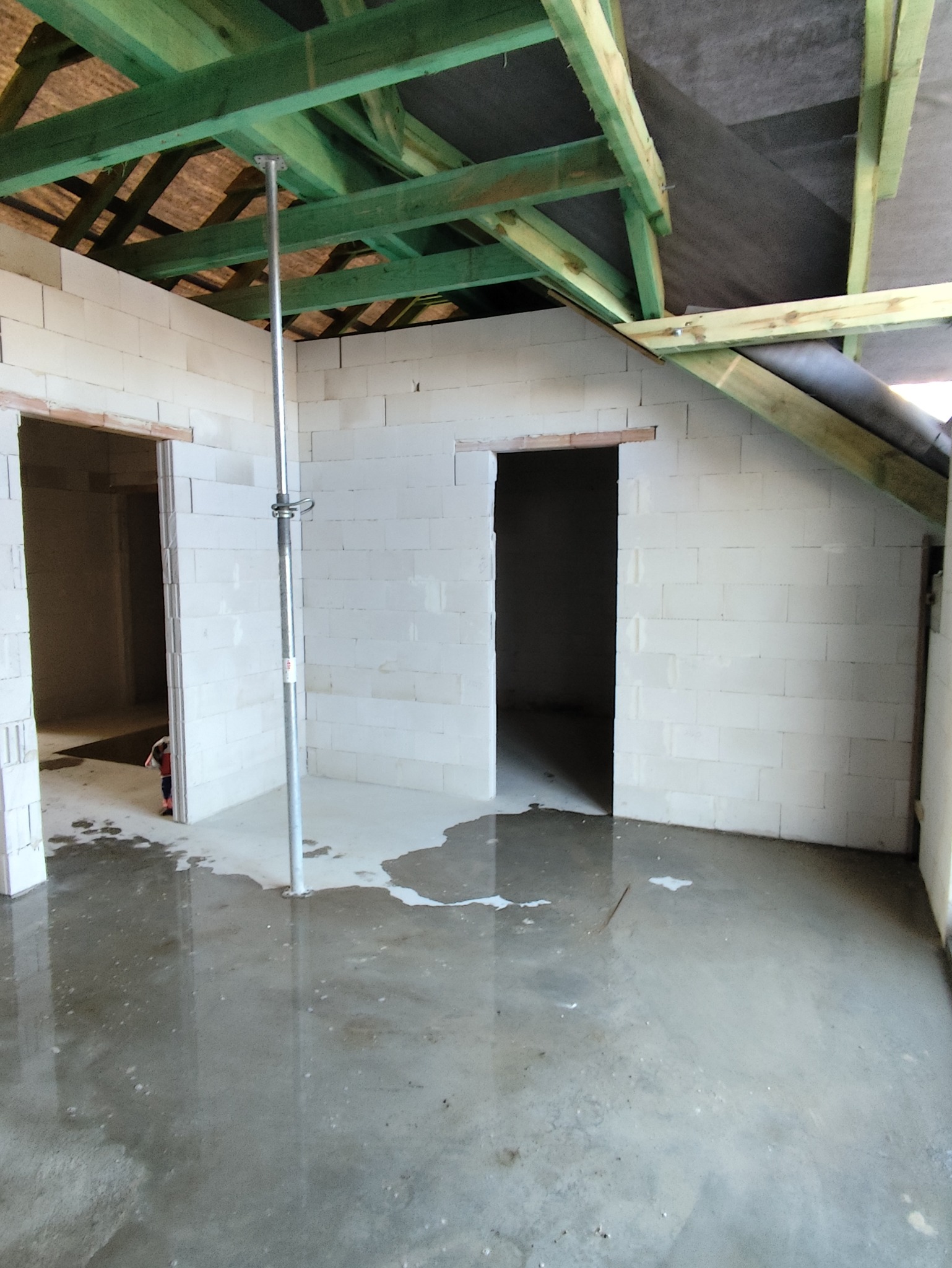 Wnętrze w trakcie murowania ścian z betonu komórkowego, widoczne drewniane elementy konstrukcji dachu i mokra posadzka. Podpora stropowa w centrum kadru.