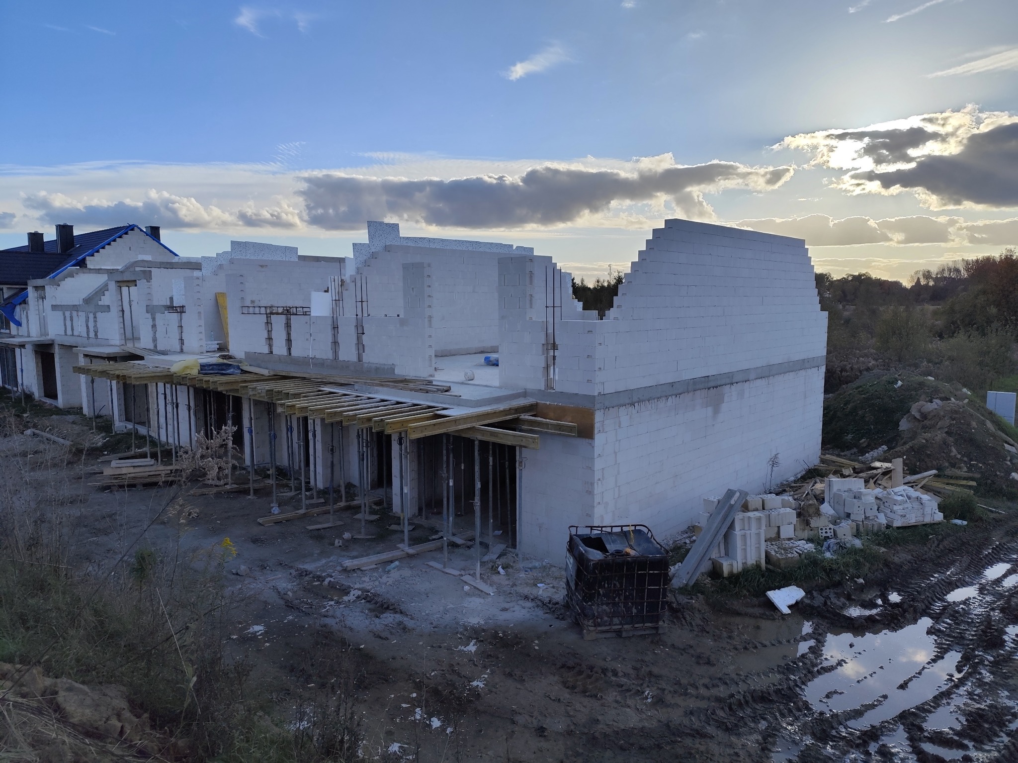 Wznoszenie ścian z białych bloczków na budowie domu jednorodzinnego, widoczne podpory stropu i materiały budowlane. W tle zachmurzone niebo.