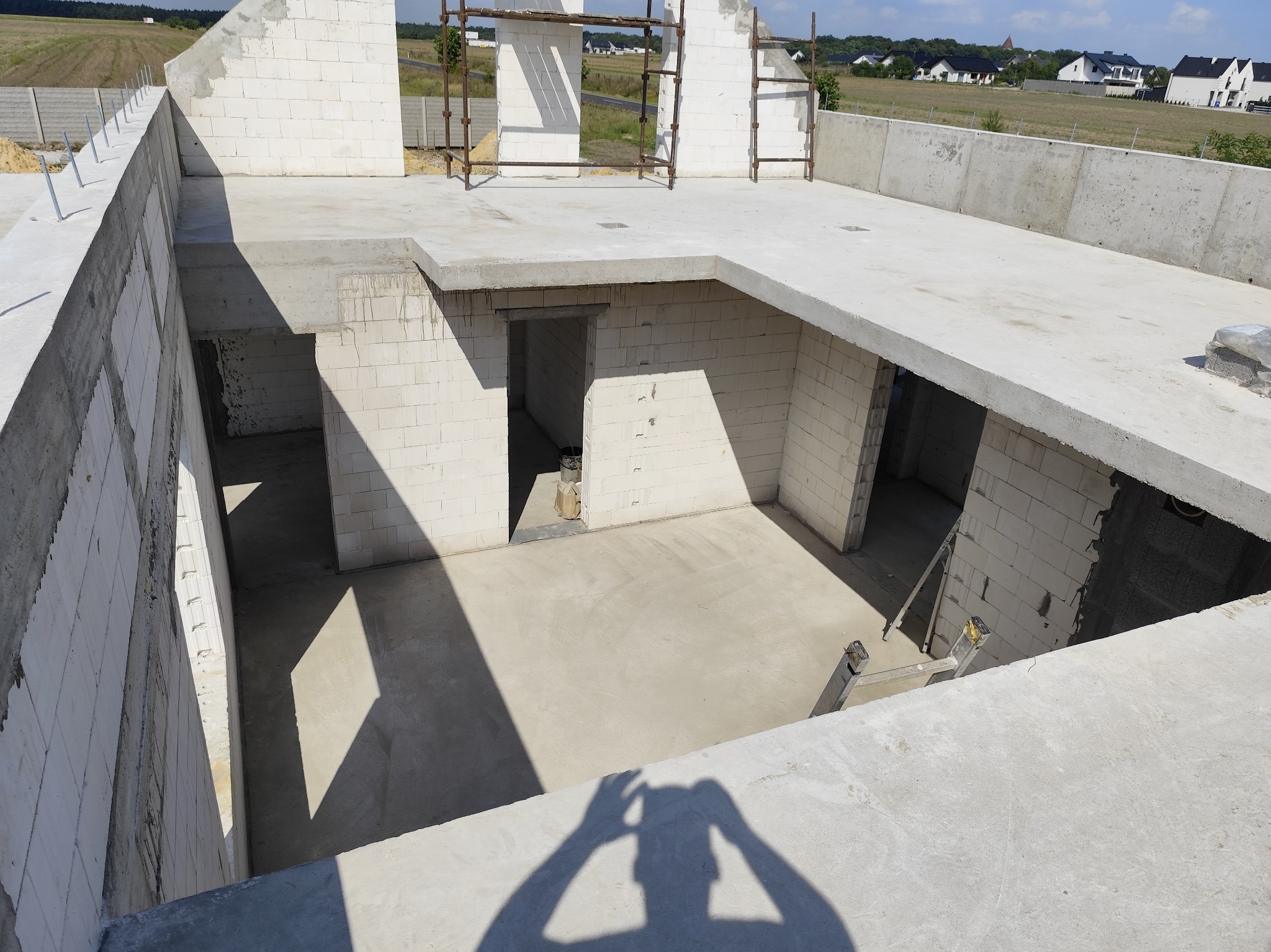 Surowy stan budynku w trakcie budowy: białe ściany z betonu komórkowego, wylany betonowy strop, widoczne otwory drzwiowe i rusztowanie. W tle pola i zabudowa jednorodzinna.