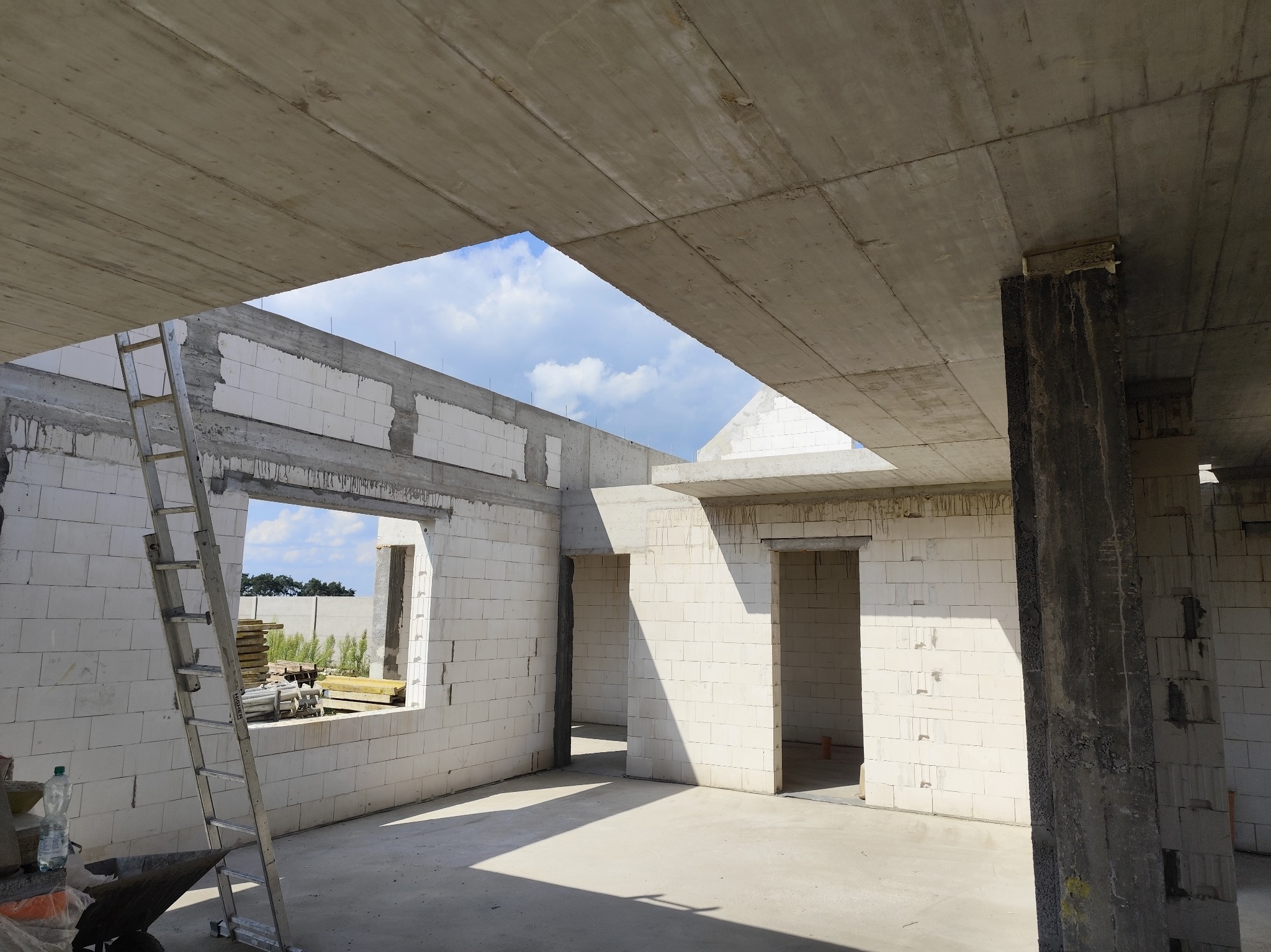 Wnętrze surowego domu w trakcie budowy z białych bloczków, widoczne betonowe stropy i filar, drabina oparta o ścianę, w tle niebo przez otwory okienne.