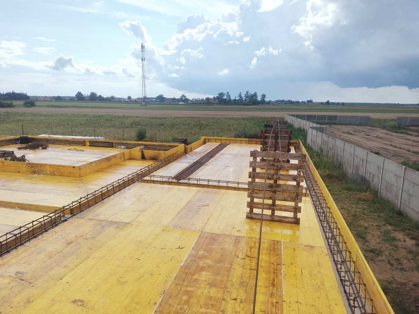 Żółte szalunki fundamentów z częściowo ułożonym zbrojeniem na tle krajobrazu z wieżą telekomunikacyjną i betonowym ogrodzeniem. Widok z góry.