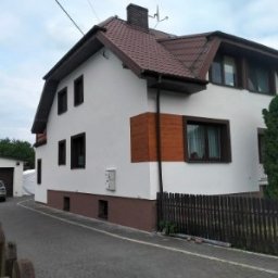 BUDEX FIRMA USŁUGOWO-HANDLOWA SPÓŁKA Z OGRANICZONĄ ODPOWIEDZIALNOŚCIĄ - Biały dom z brązowym dachem i drewnianymi akcentami, widoczny z boku, z garażem w tle i drewnianym ogrodzeniem na pierwszym planie. Widoczne okna i elewacja.