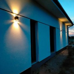 BUDEX FIRMA USŁUGOWO-HANDLOWA SPÓŁKA Z OGRANICZONĄ ODPOWIEDZIALNOŚCIĄ - Oświetlona elewacja domu z zamontowanymi lampami zewnętrznymi, widok z boku o zmierzchu. Świeżo otynkowana ściana w kolorze jasnego błękitu.