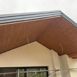 Drewniana podbitka dachowa z szarym okapem w nowoczesnym domu. Widoczne fragmenty rusztowania i wystające przewody. Jasna elewacja budynku.