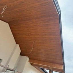 Drewniana podbitka dachowa z widocznymi elementami konstrukcyjnymi i fragmentem rusztowania. Ujęcie z dołu, podkreślające strukturę drewna i detale wykończenia.