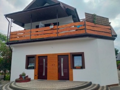 Dwupiętrowy dom z białymi ścianami i drewnianymi elementami wykończeniowymi, balkon z oryginalną balustradą. Widoczne drzwi wejściowe i okna z kwiatami.