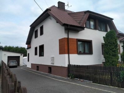 Biały dom z brązowym dachem i drewnianymi akcentami, widoczny z boku, z garażem w tle i drewnianym ogrodzeniem na pierwszym planie. Widoczne okna i elewacja.