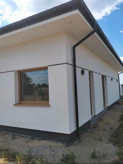 Biała elewacja domu jednorodzinnego z drewnianą stolarką okienną i czarnymi rynnami, widok z zewnątrz w słoneczny dzień. Wykończona podmurówka w kolorze grafitowym.