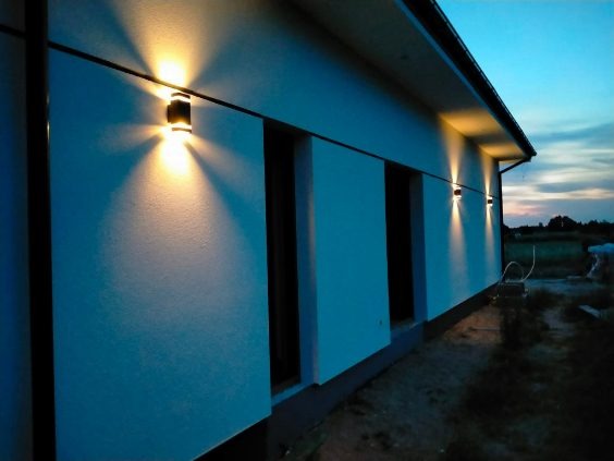 Oświetlona elewacja domu z zamontowanymi lampami zewnętrznymi, widok z boku o zmierzchu. Świeżo otynkowana ściana w kolorze jasnego błękitu.