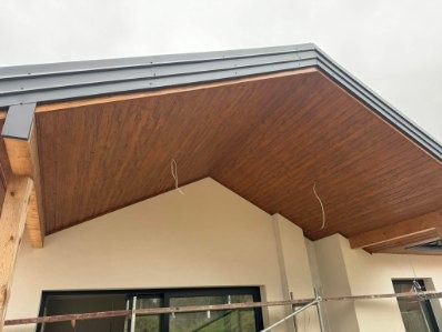 Drewniana podbitka dachowa z szarym okapem w nowoczesnym domu. Widoczne fragmenty rusztowania i wystające przewody. Jasna elewacja budynku.