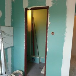 Build-Master Marcin Wziontek - Wnętrze w trakcie prac: ściany z płyt gipsowo-kartonowych, częściowo wyszpachlowane, widoczne narzędzia i wiadro z masą szpachlową, przygotowanie do dalszych prac wykończeniowych.