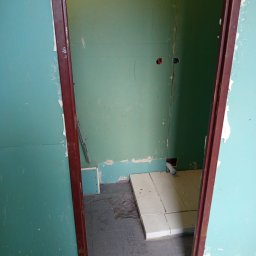 Build-Master - Łazienka w trakcie remontu: zielone płyty gipsowe, częściowo ułożone białe płytki podłogowe, widoczne instalacje. Ciemna futryna drzwi.