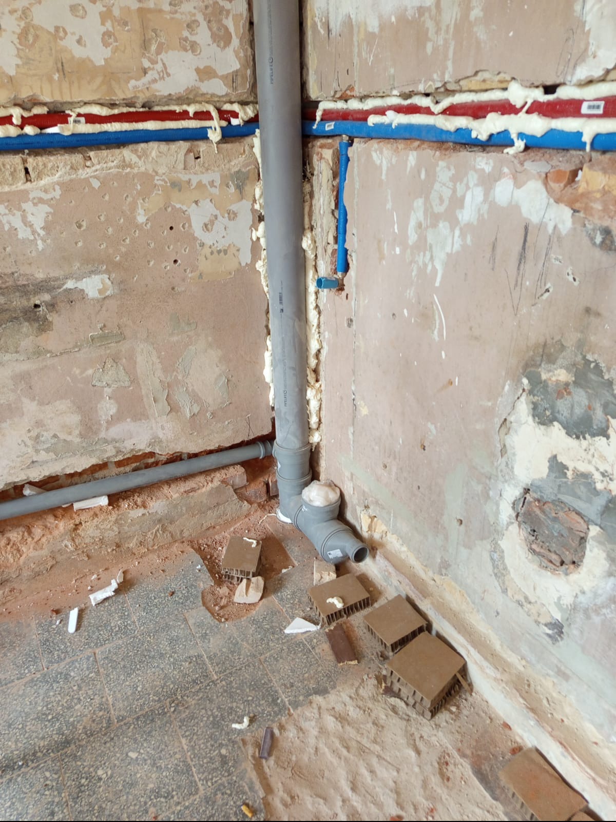 Szare rury kanalizacyjne połączone kolankiem, prowadzone wzdłuż ściany z widocznymi instalacjami wodnymi zabezpieczonymi pianką montażową. Fragment podłogi z płytek.