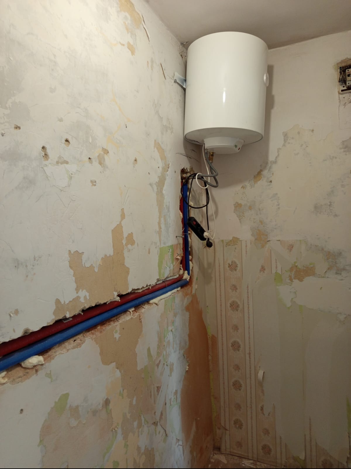 Bojler elektryczny zamontowany na ścianie w łazience w trakcie remontu. Widoczne rury instalacji wodnej i fragmenty starej tapety.