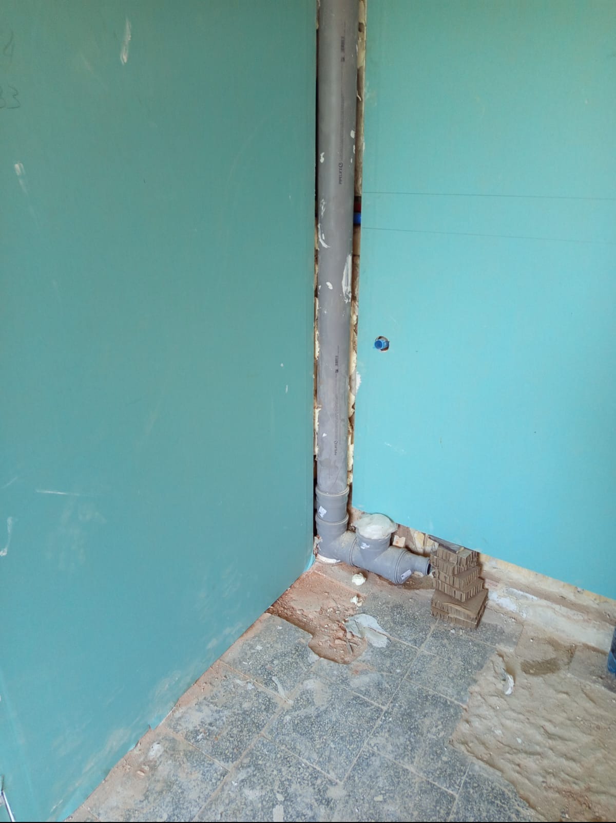 Pionowa rura kanalizacyjna w rogu pomieszczenia z płytami kartonowo-gipsowymi, widoczne łączenia i pianka montażowa. Fragment podłogi z szarych płytek.