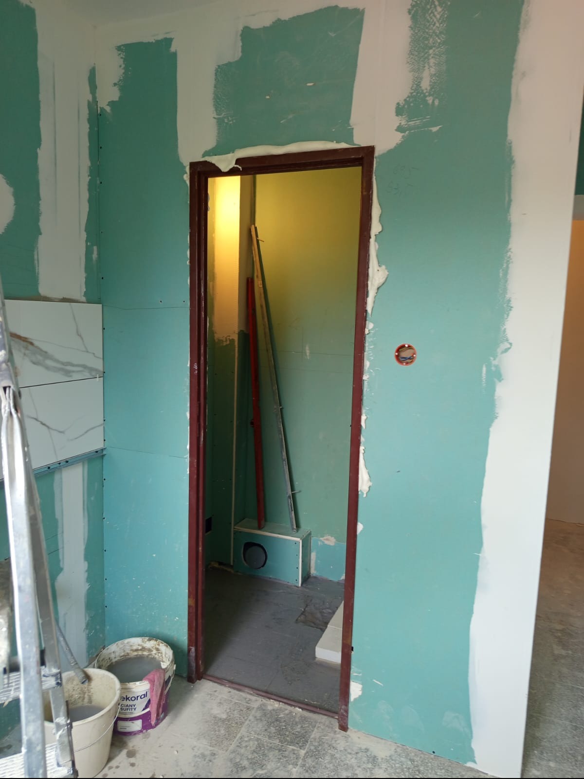 Wnętrze w trakcie prac: ściany z płyt gipsowo-kartonowych, częściowo wyszpachlowane, widoczne narzędzia i wiadro z masą szpachlową, przygotowanie do dalszych prac wykończeniowych.