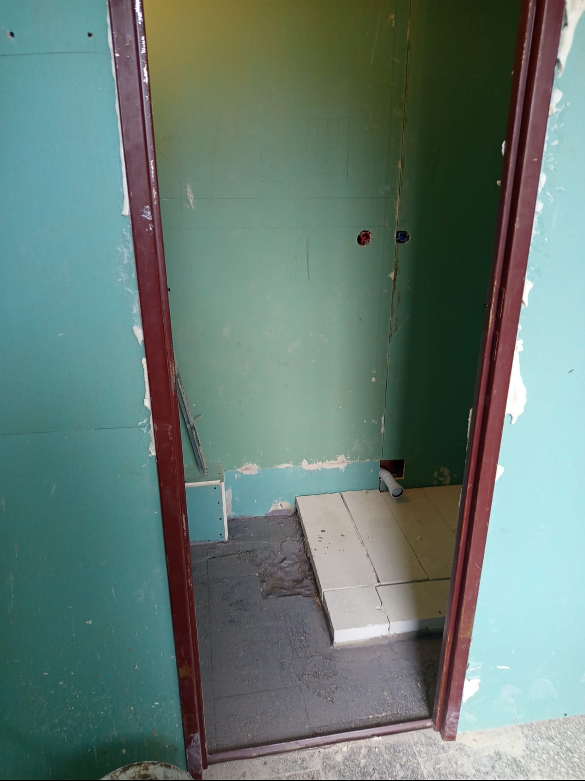 Łazienka w trakcie remontu: zielone płyty gipsowe, częściowo ułożone białe płytki podłogowe, widoczne instalacje. Ciemna futryna drzwi.