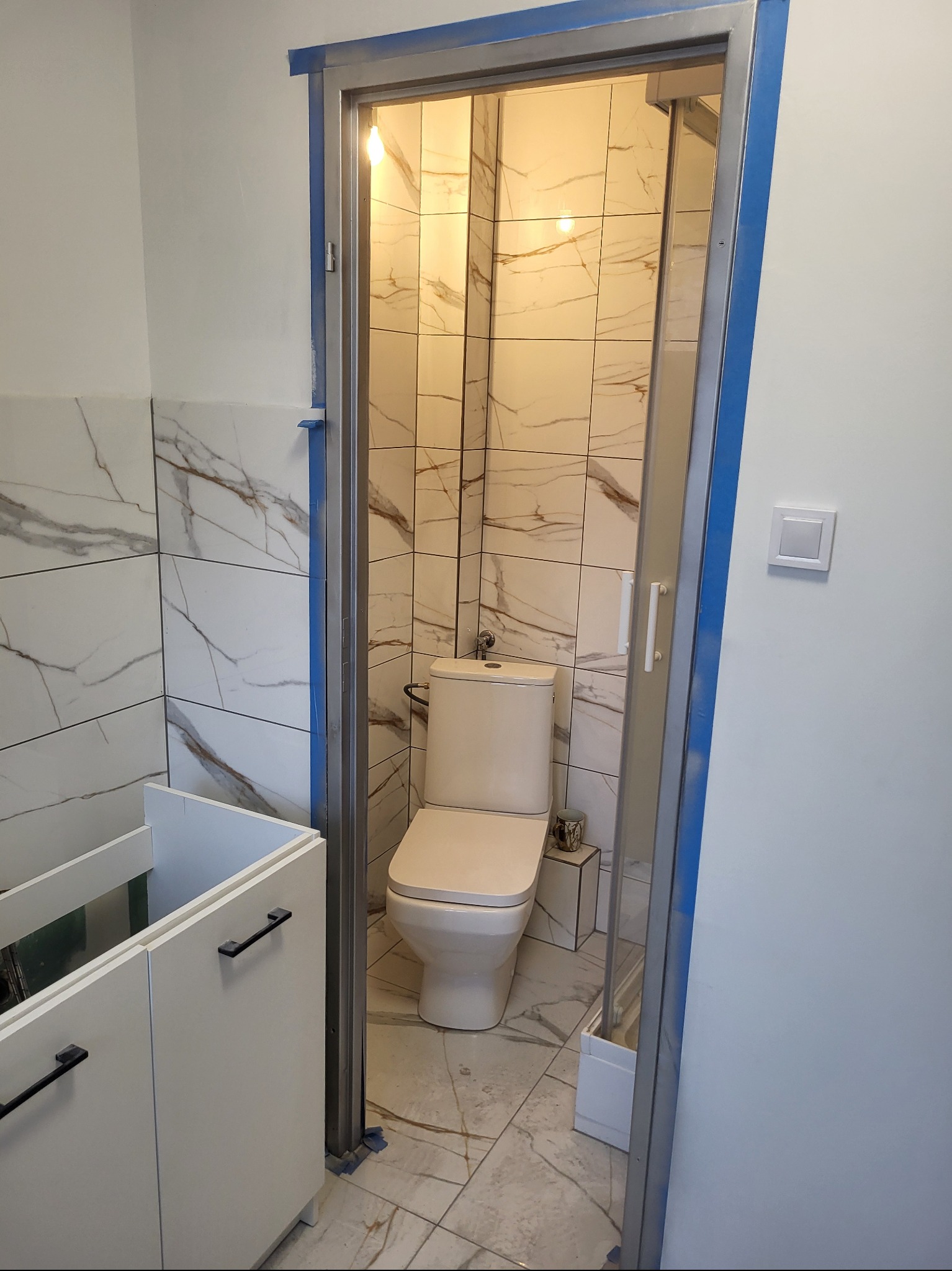 Wykończona łazienka z marmurowymi płytkami, widok przez metalową futrynę. Biała toaleta i szafka pod umywalkę z czarnymi uchwytami. Nowoczesne wnętrze po remoncie.