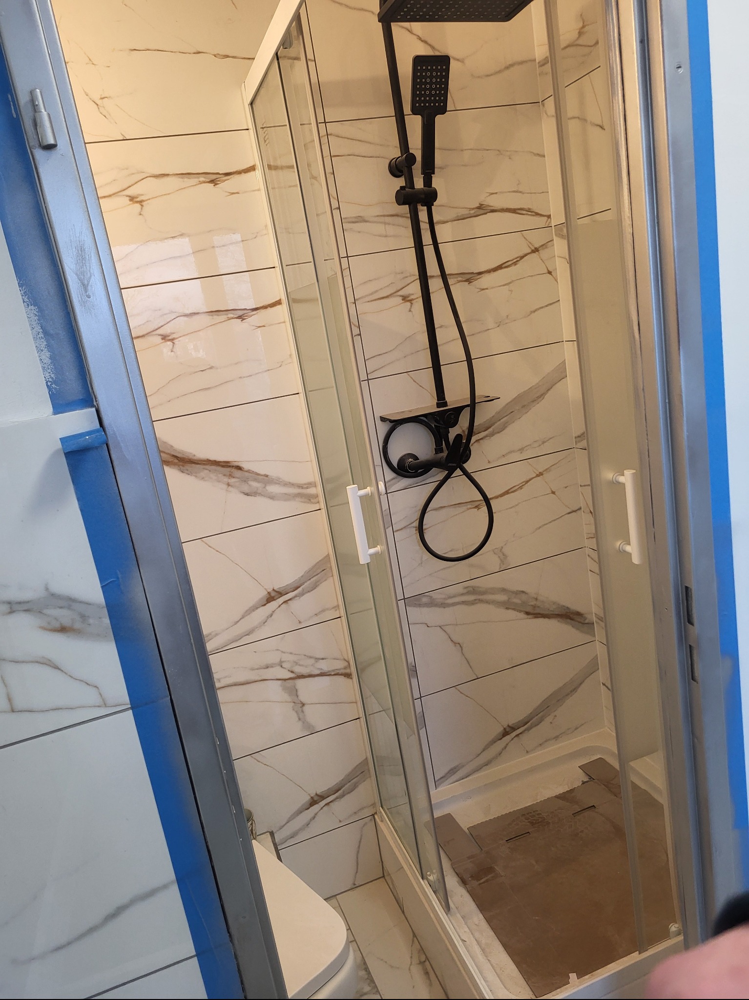 Kabina prysznicowa z czarną armaturą na tle płytek imitujących marmur. Widoczne detale wykończenia i zabezpieczenia folią malarską.