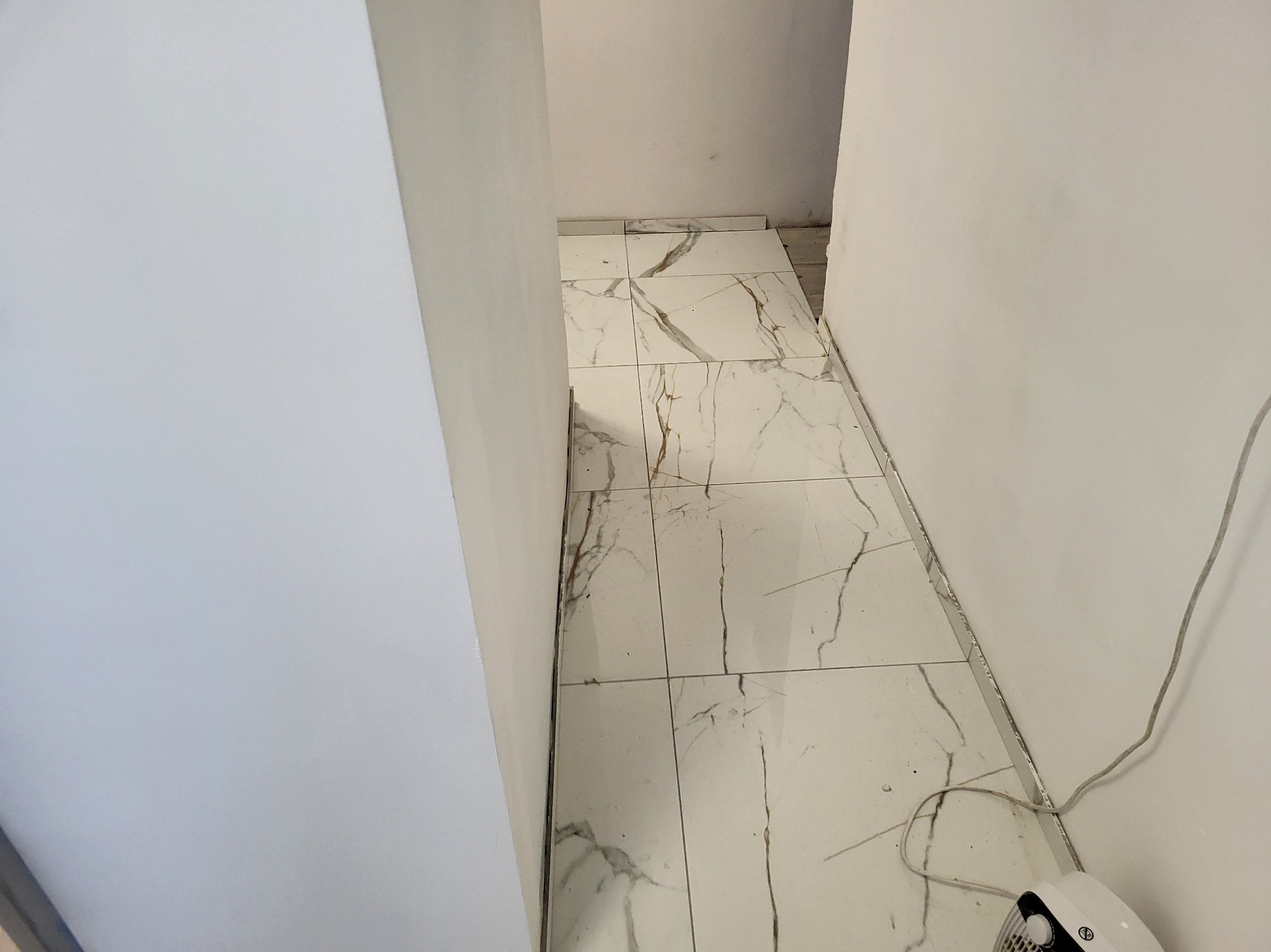 Ułożona podłoga z białych płytek imitujących marmur w wąskim korytarzu. Widoczne białe ściany i fragment urządzenia elektrycznego przy podłodze.