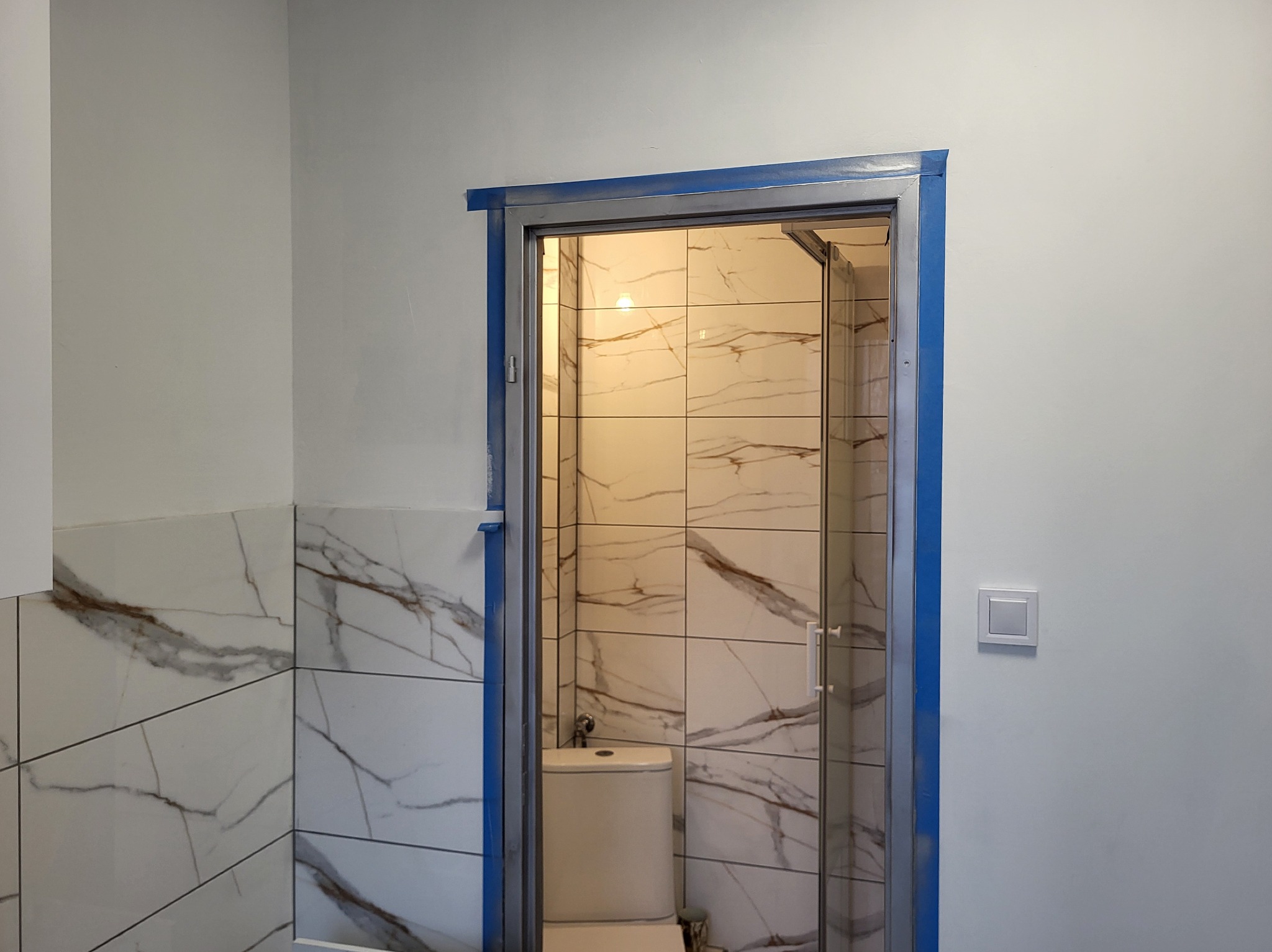 Wykończona łazienka z marmurowymi płytkami i kabiną prysznicową za szklanymi drzwiami. Widoczny fragment toalety. Świeżo pomalowane ściany i nowy włącznik światła.